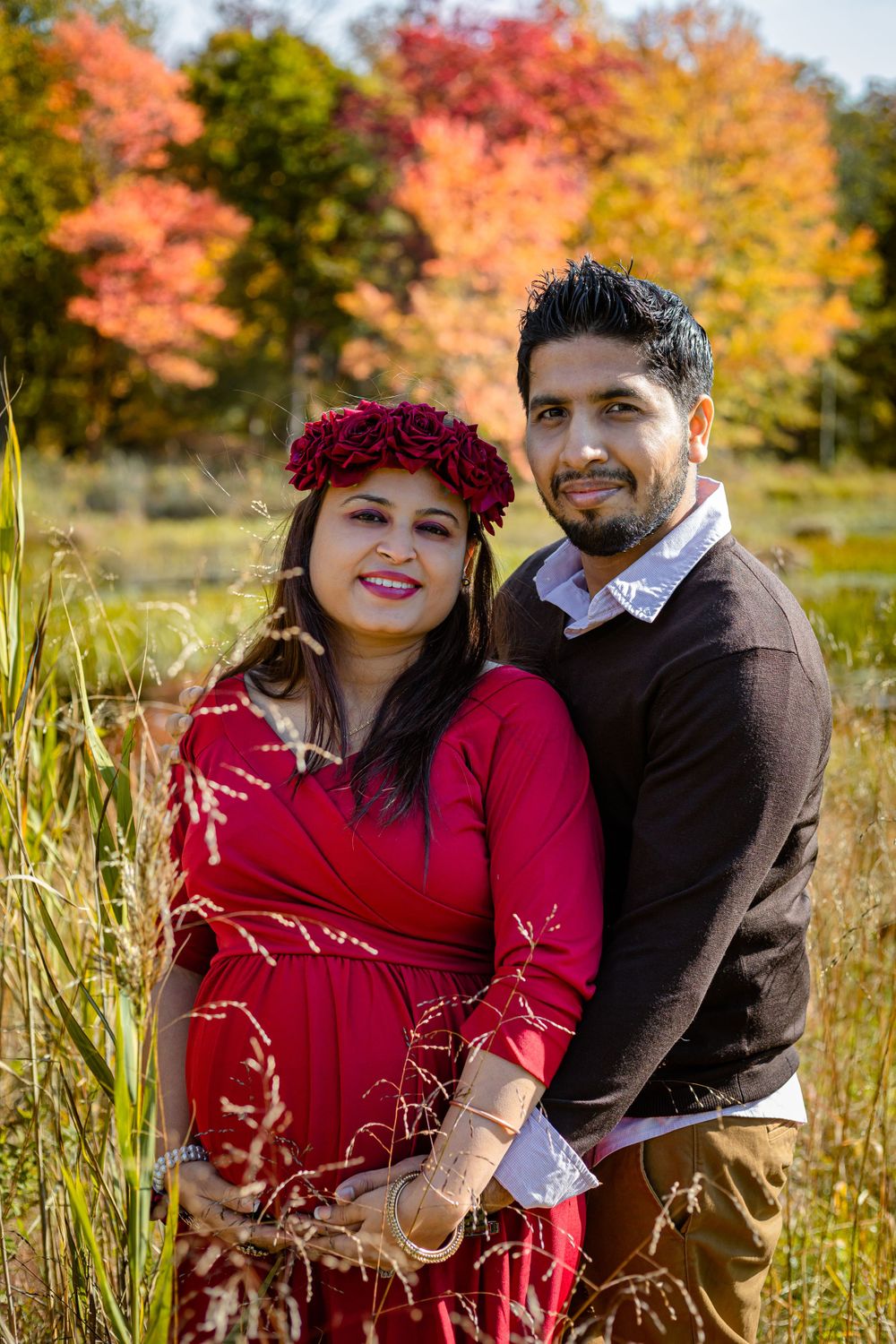 A couple poses in a field with vibrant autumn foliage, the woman wearing a red dress and floral crown.