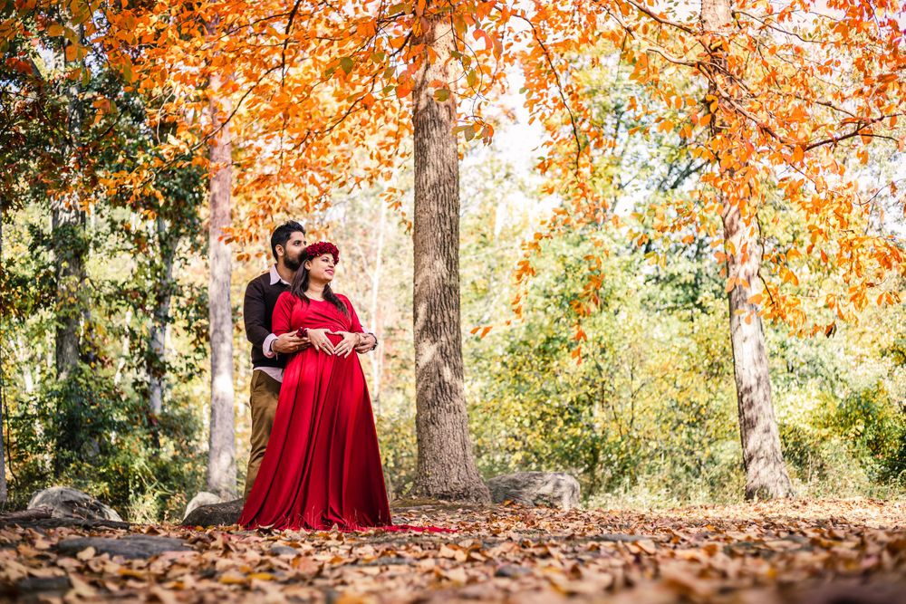 A couple stands among fallen leaves beneath orange autumn trees, with the woman in a flowing red gown.