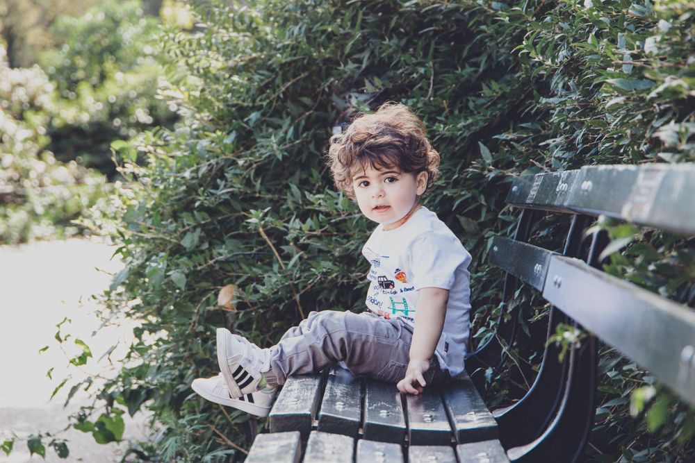 A small child sits alone on a wooden park bench surrounded by greenery.