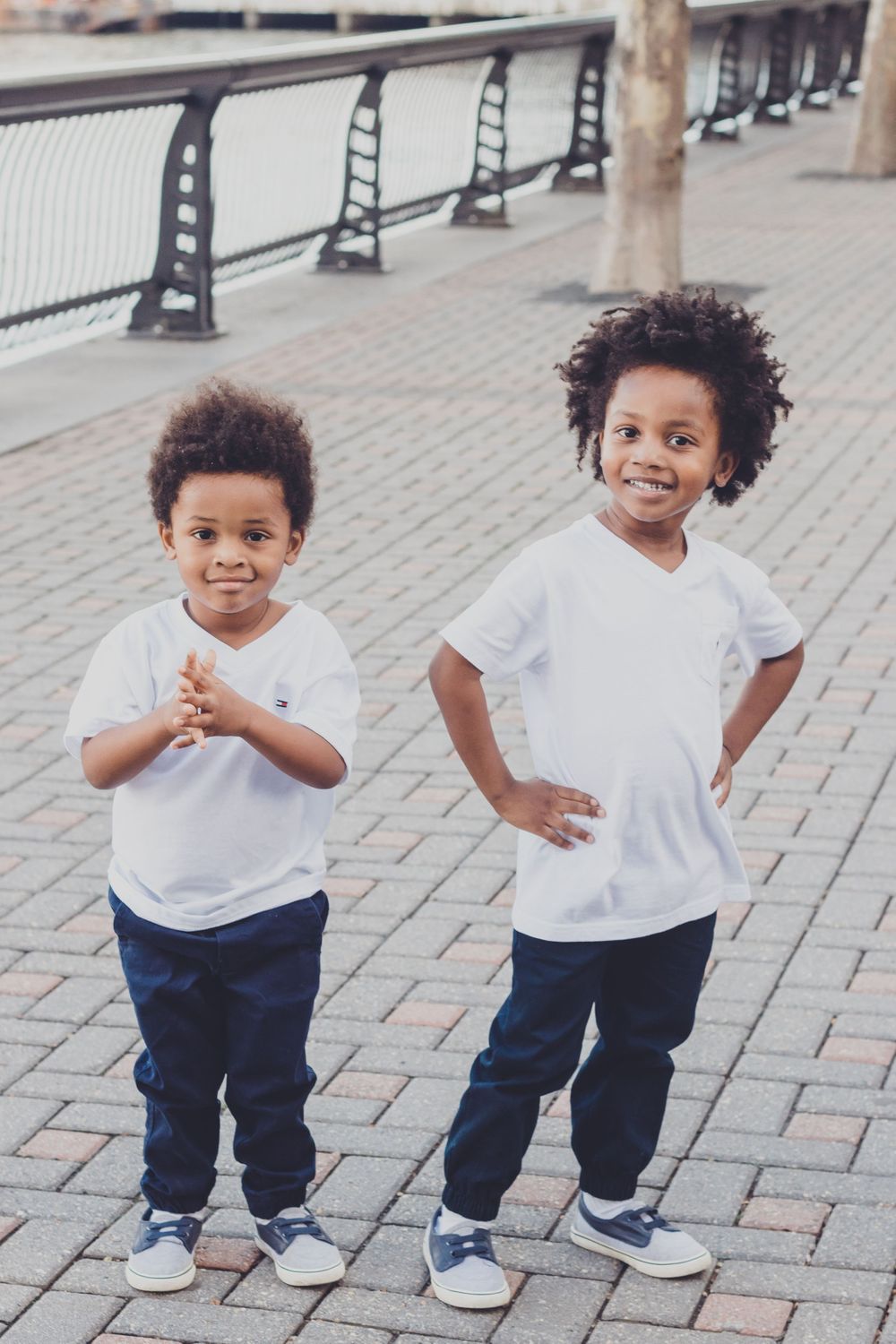 Playful toddler in white t-shirt and blue pants makes expressive poses on brick pavement.
