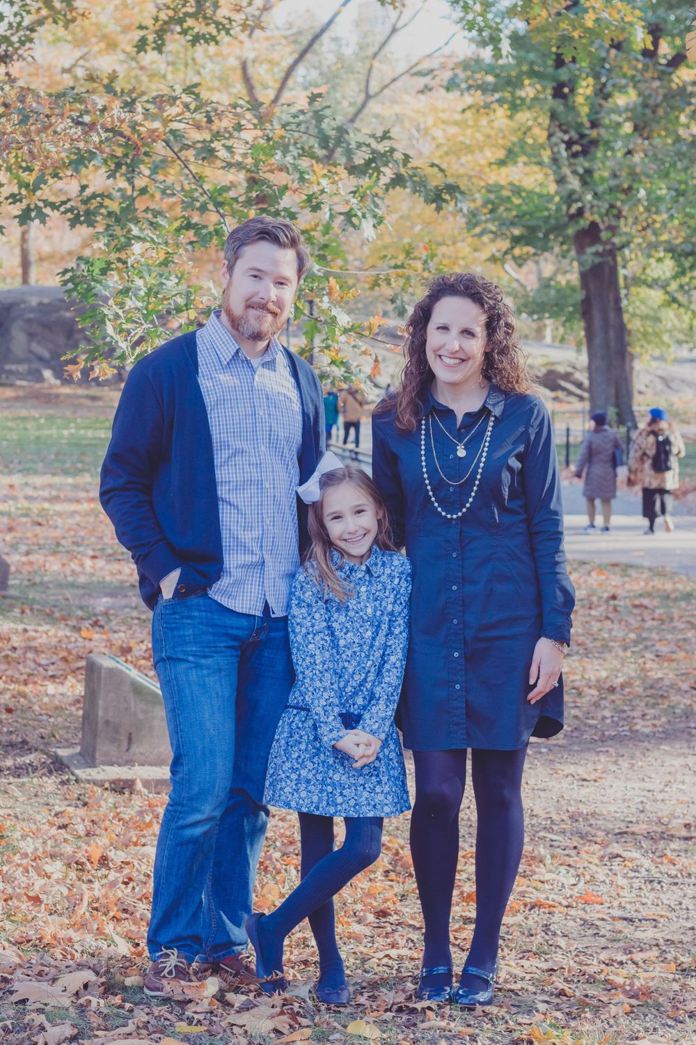 Family of three posing together in casual outfits among autumn trees.