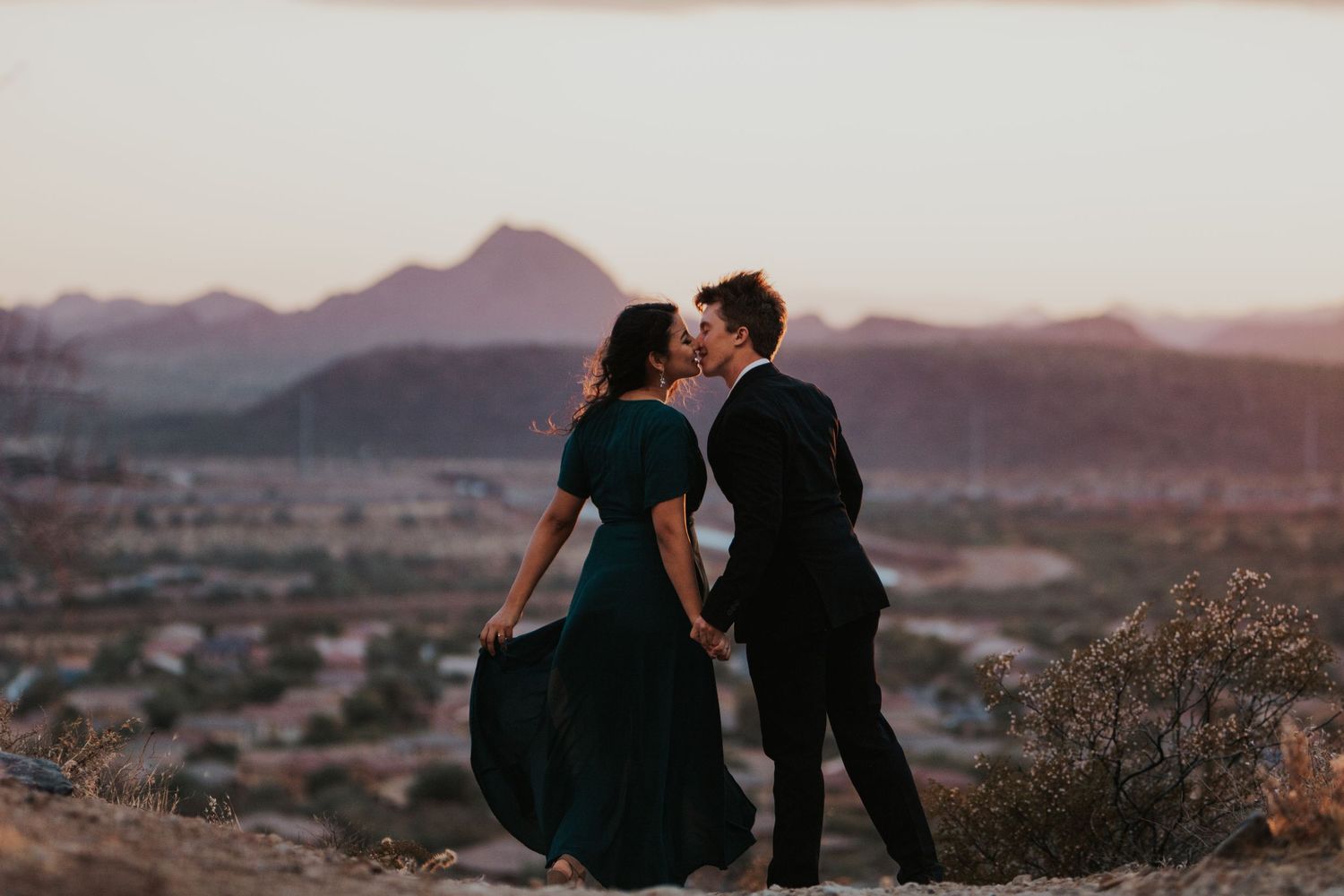 Engaged couple inches from kissing in a dress and suit overlooking a desert landscape at sunset.