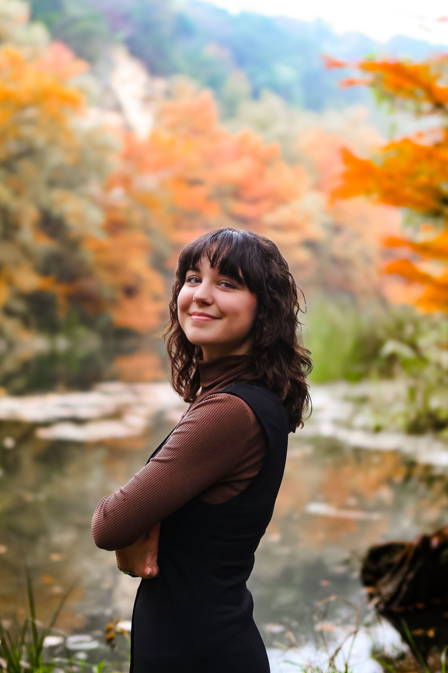 A person in dark clothing poses against vibrant autumn foliage near a stream.