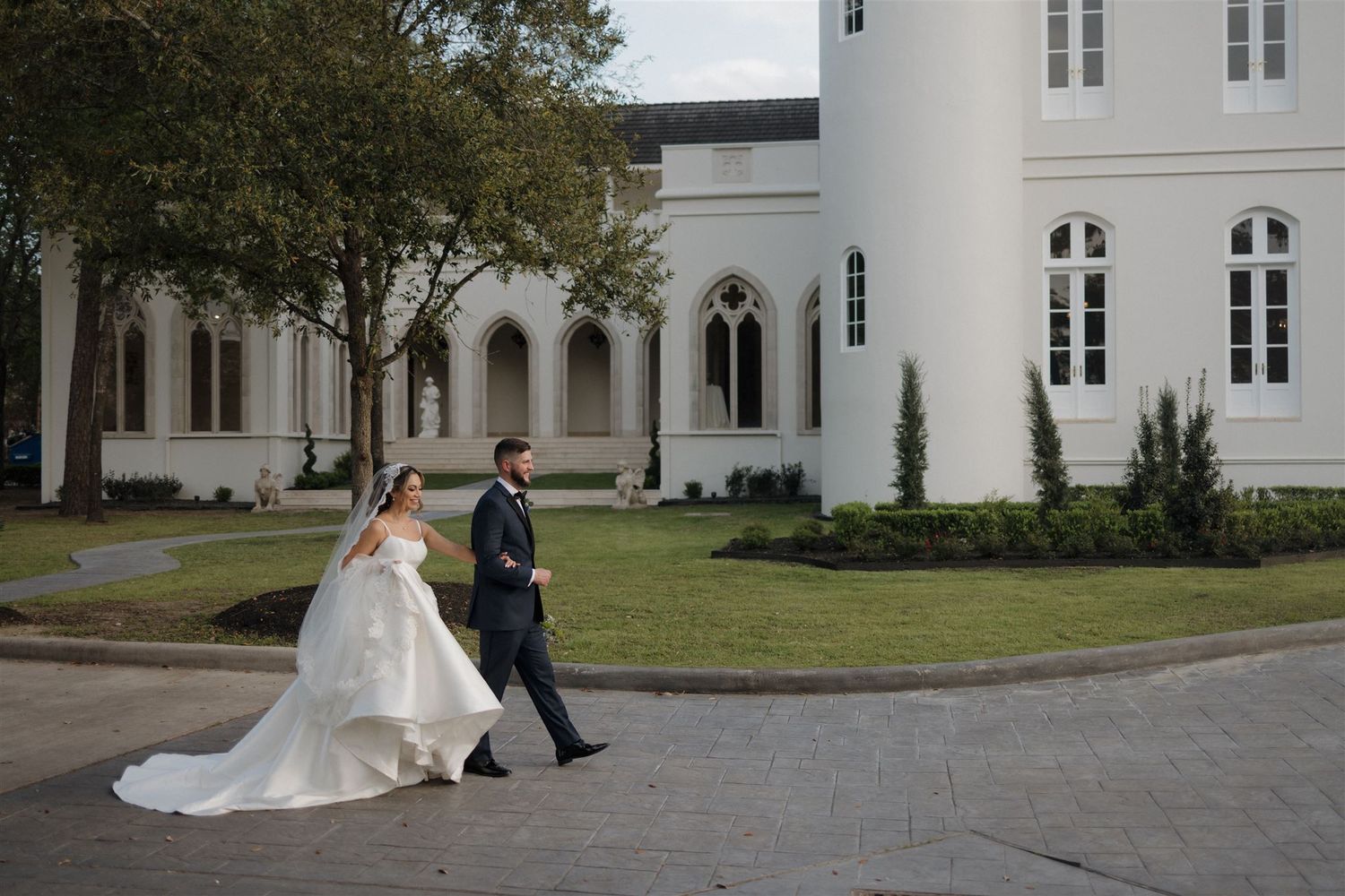 Couple strolls along curved driveway outside white gothic-style church building.