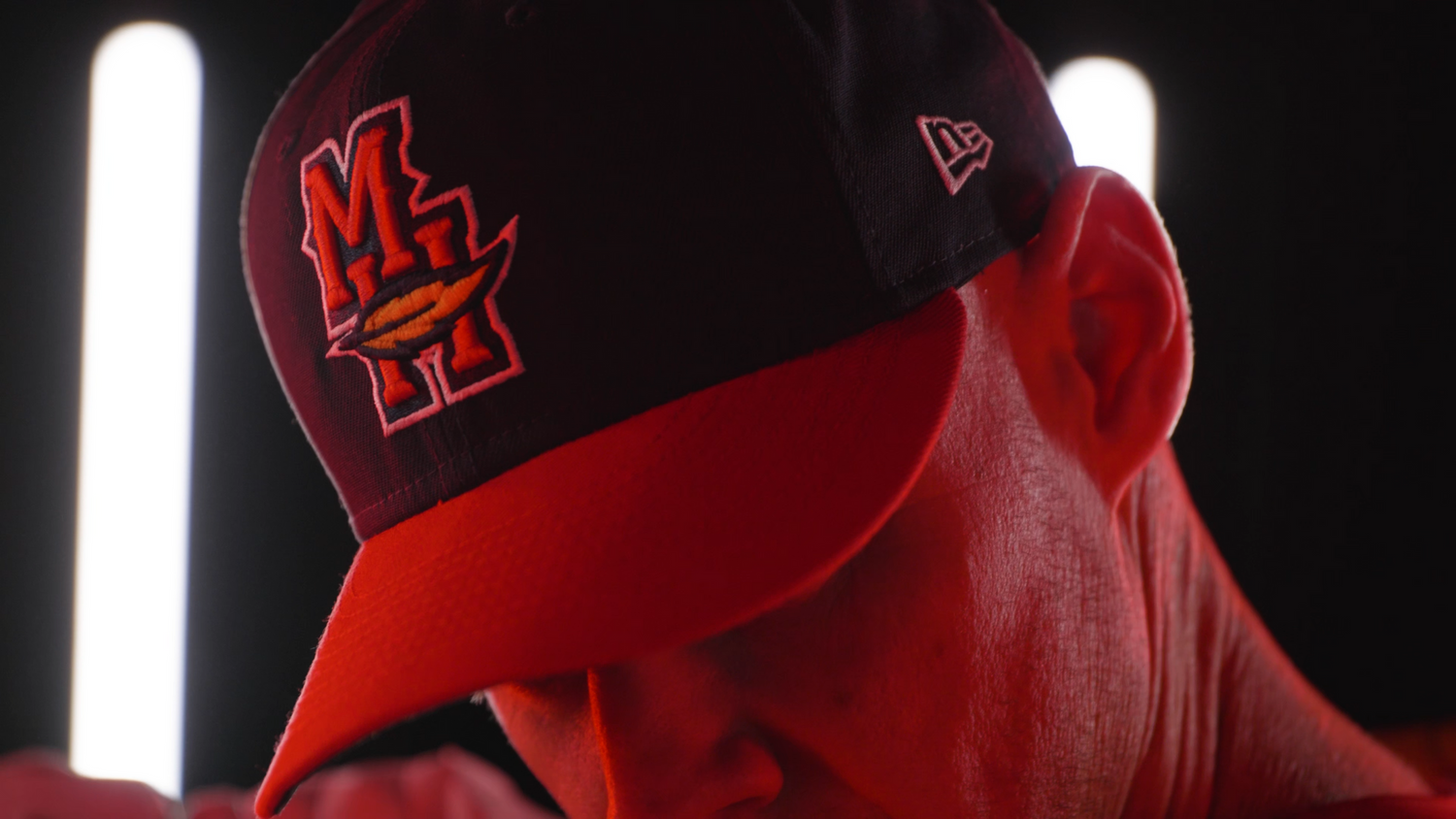 Close-up of red baseball cap with dark logo against black background with light pillars.