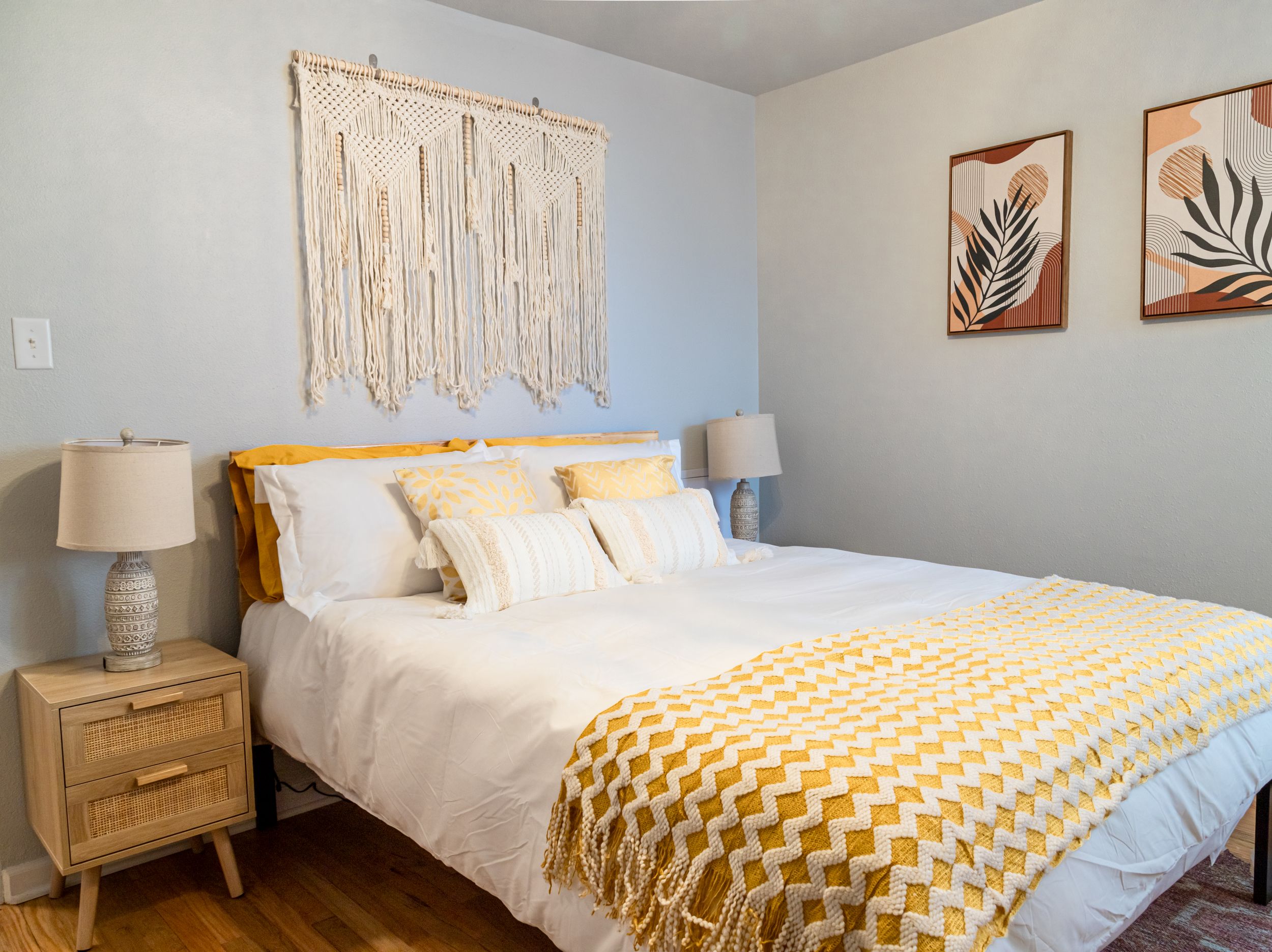 Modern bedroom with macrame wall hanging, yellow throw blanket, and botanical art prints.