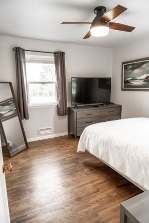 Bedroom with ceiling fan, window with curtains, mounted TV, and landscape artwork on gray walls.