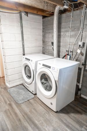 Two white washing machines set up in a basement laundry area with concrete walls and wood flooring.