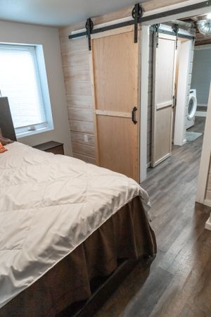 Modern bedroom with sliding barn door and white bedding features wood flooring and window lighting.