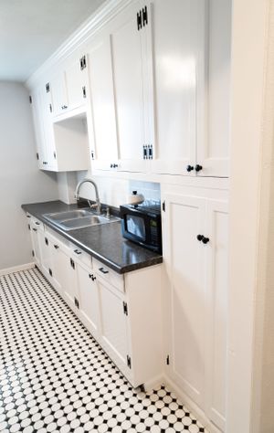 Bright galley kitchen with white shaker cabinets, dark countertops, and black and white checkerboard flooring.