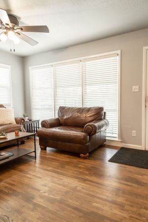 Living room with leather armchair, ceiling fan, and large windows with white blinds.