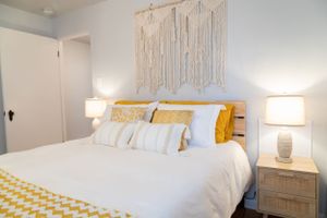 Modern bedroom with white bedding, mustard yellow accents, and natural macrame wall art.