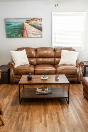 Cozy living room with brown leather couch, coastal artwork, and modern coffee table on hardwood floors.