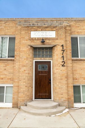 Row of brown brick apartment units with wooden doors and unit numbers along exterior wall.