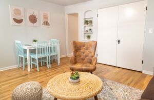 Cozy living area featuring woven coffee table, tufted armchair, and mint green dining set with abstract wall art.