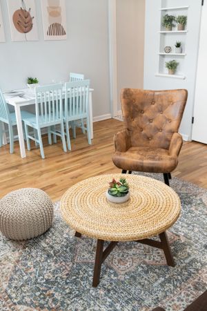 Modern living space featuring a round woven coffee table, tan leather chair, and knit pouf on patterned rug.