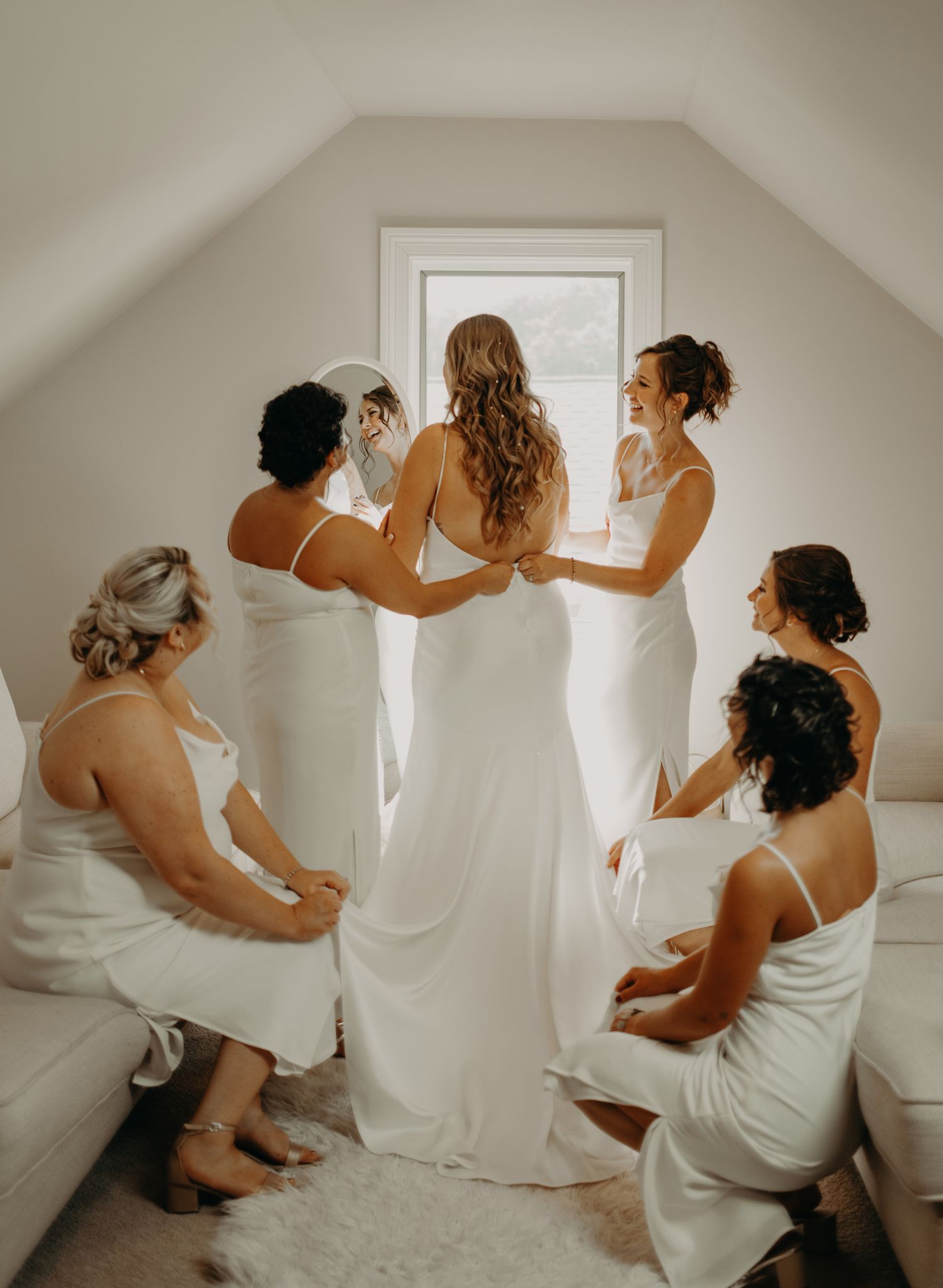 Wedding party in white dresses helping with final dress preparations in a bright room.