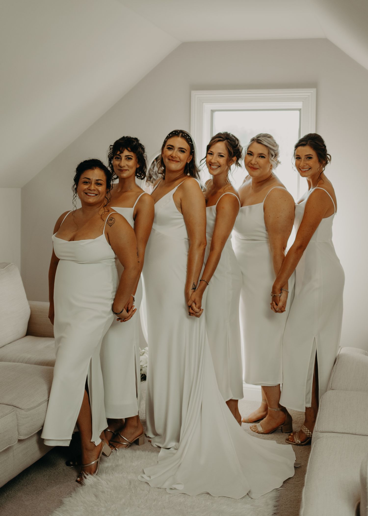 Bridal party in matching white dresses posing together in a bright attic room.