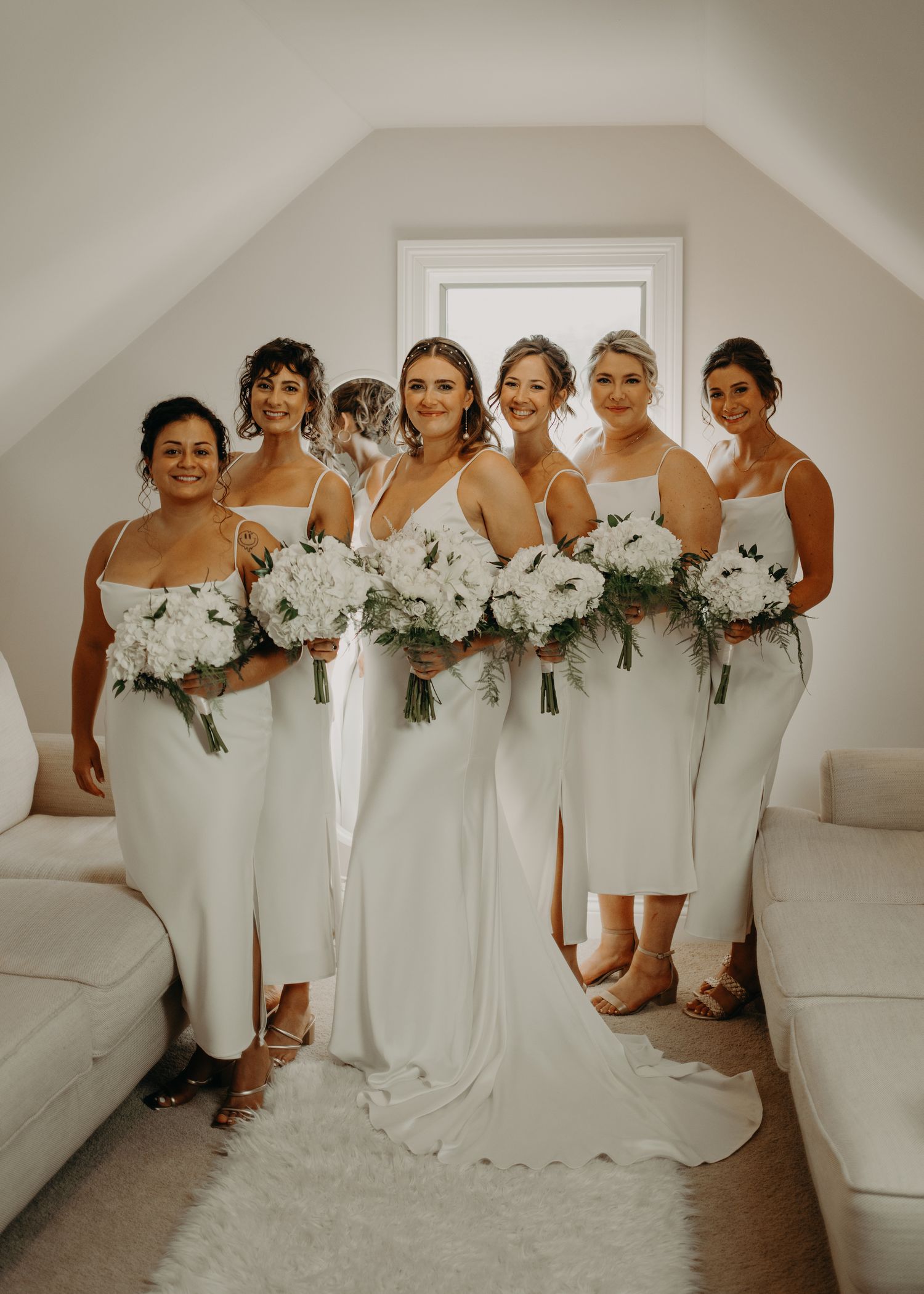Bridal party holding white bouquets pose together in elegant white dresses.