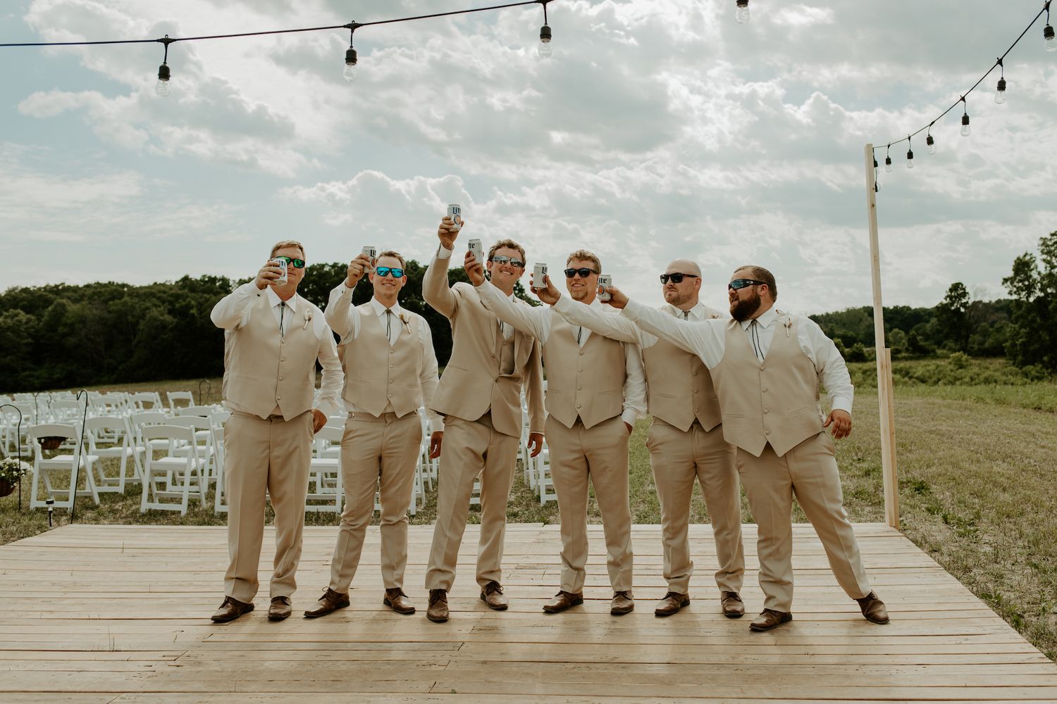 Groomsmen in khaki suits celebrating on wooden deck under string lights.