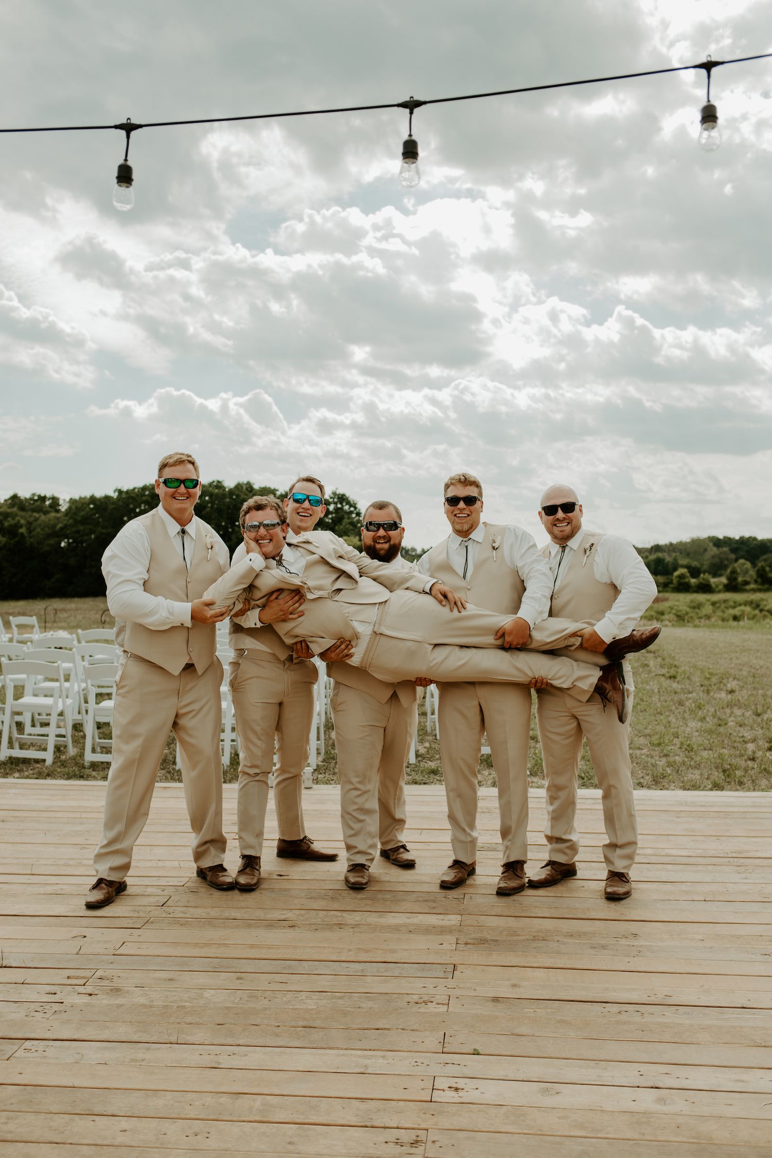 Groomsmen in matching tan suits playfully lifting someone on an outdoor wooden deck.