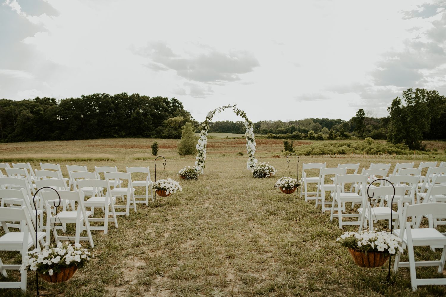 Outdoor wedding ceremony setup with white arch and chairs on a grassy field.