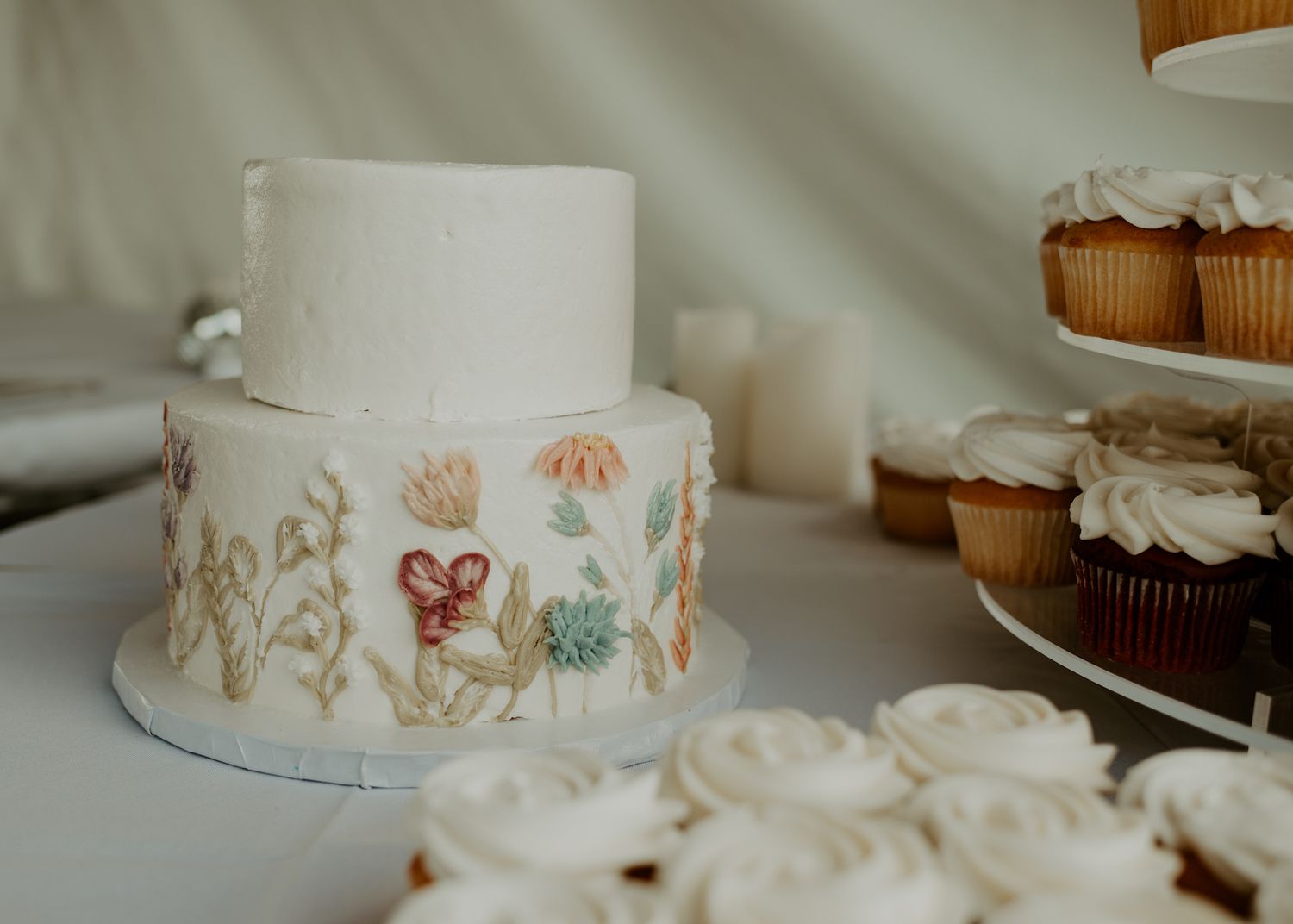 White wedding cake with delicate floral designs and cupcakes on display.