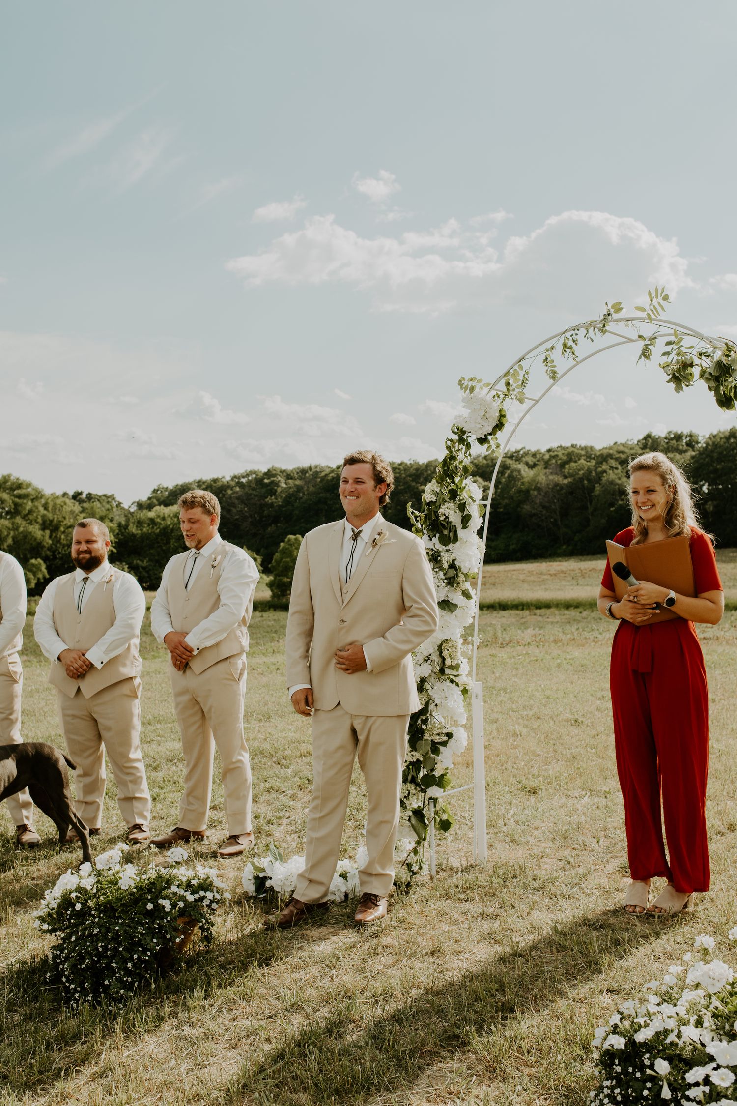 Outdoor wedding ceremony with floral arch and attendants in light suits and red attire.