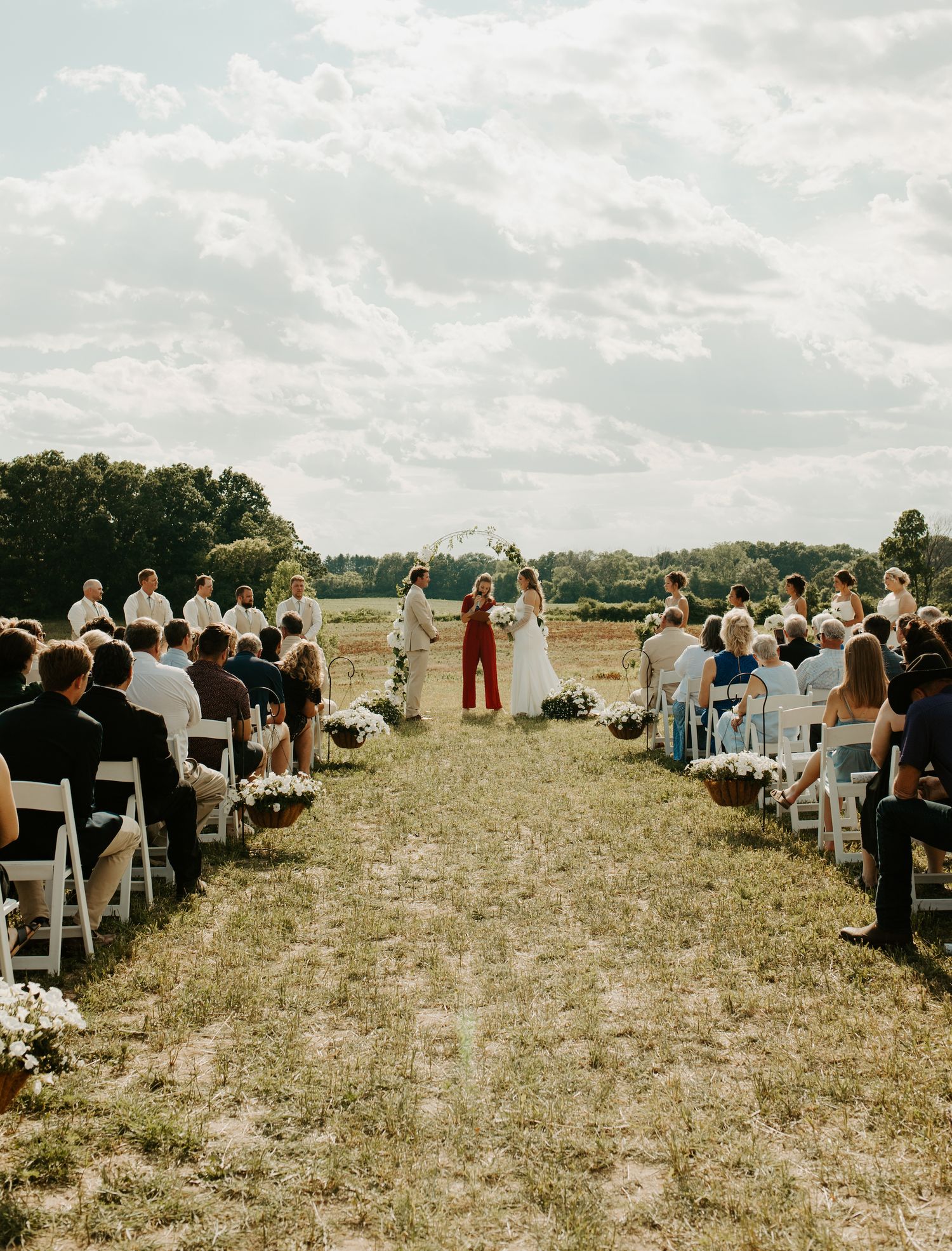 Outdoor wedding ceremony with guests seated in white chairs on a grassy field.