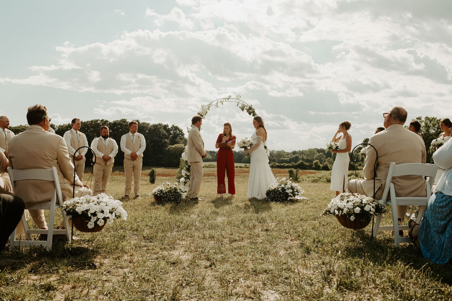 Wedding ceremony under blue sky with floral arch and guests seated on white chairs.