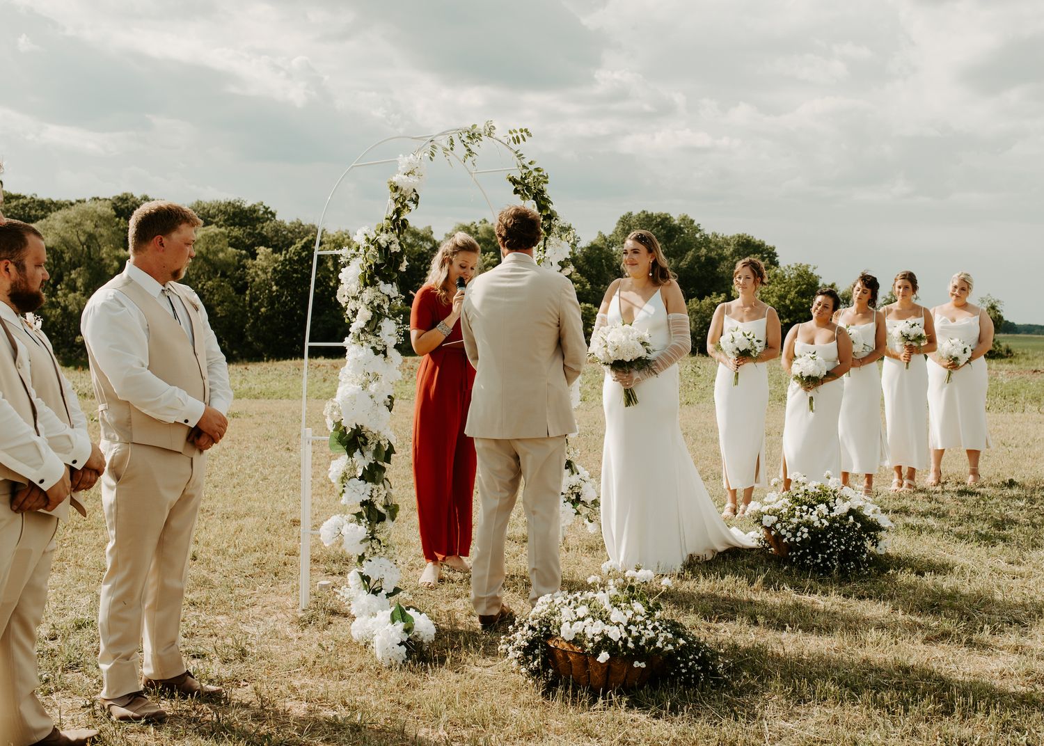 Outdoor wedding ceremony setting with floral arch and wedding party in neutral colors.