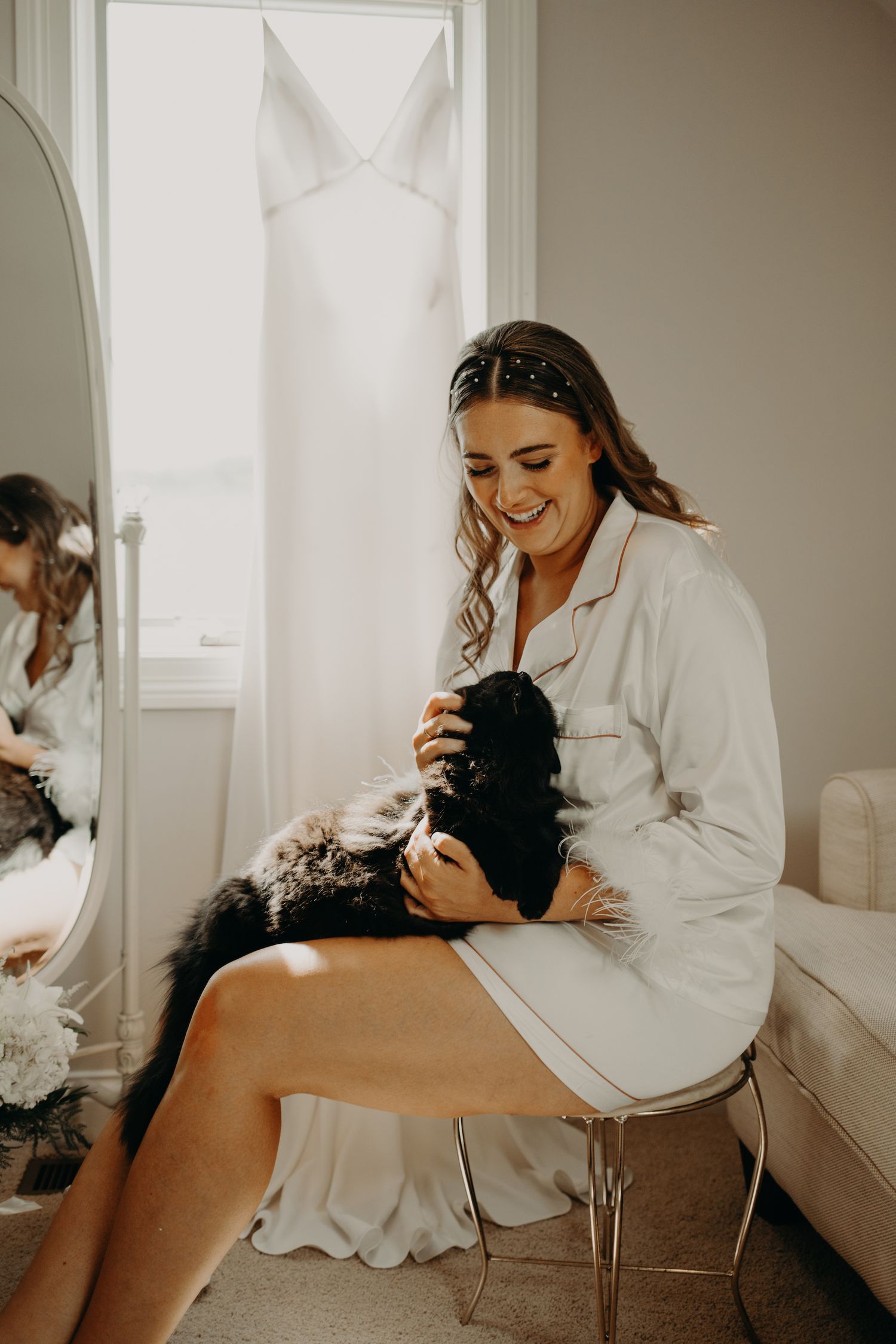 Person in white silk robe holds small black dog while sitting in bridal getting ready room.