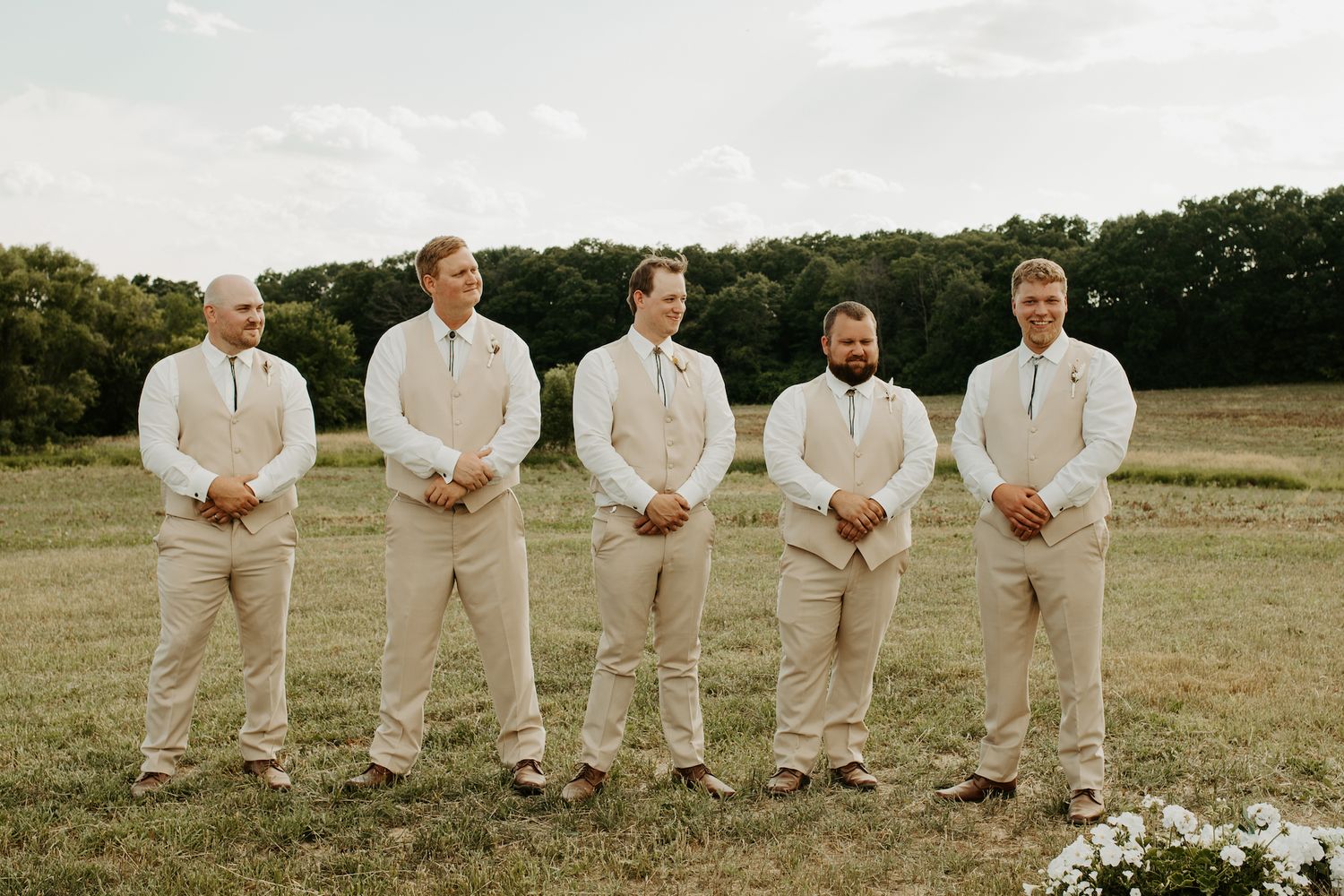 Groomsmen in beige suits standing in line on grass field.