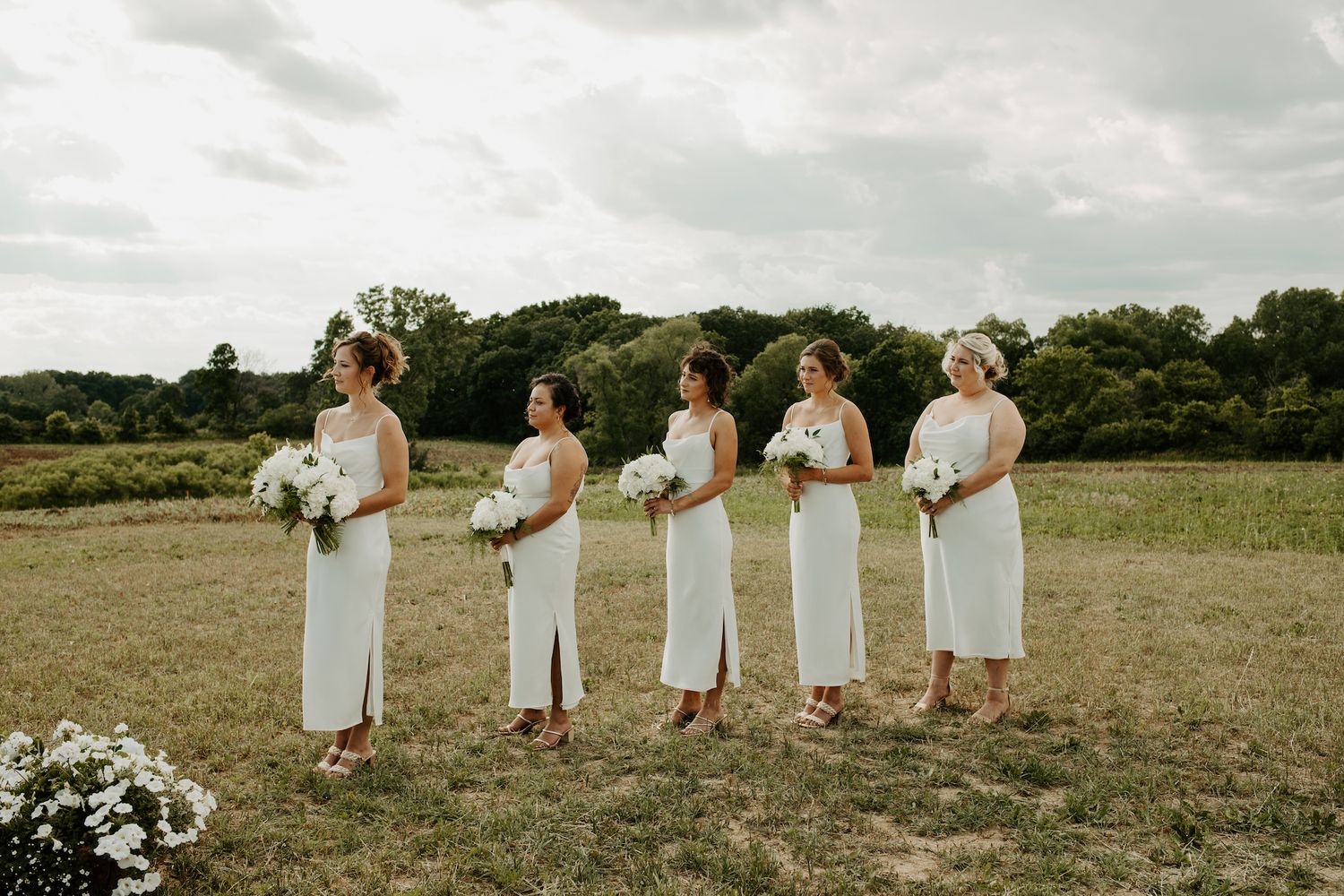 Five bridesmaids in elegant white dresses stand in a row holding bouquets in a rustic outdoor field setting.