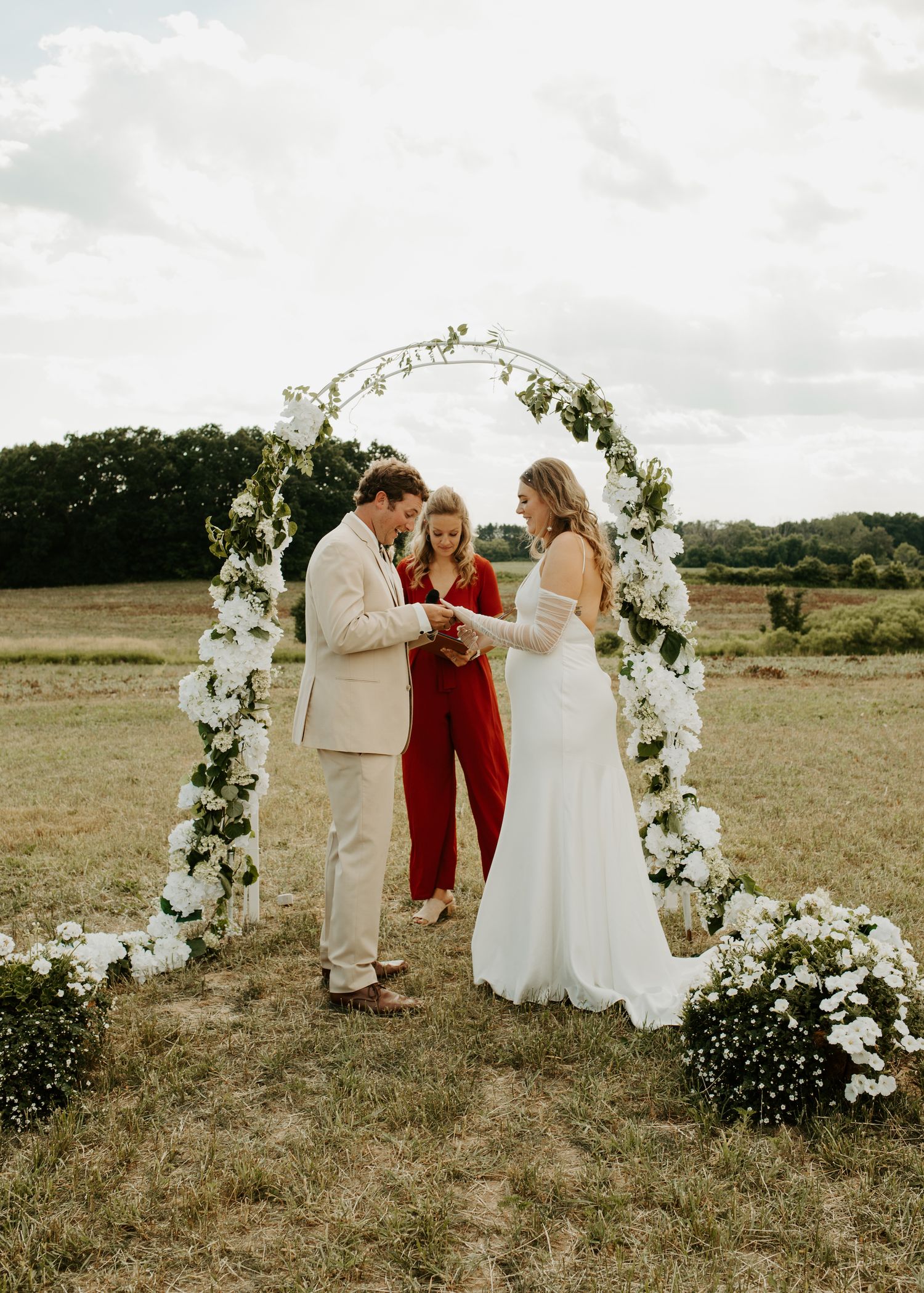 Wedding ceremony taking place under an elegant white floral arch with loved ones gathered around.