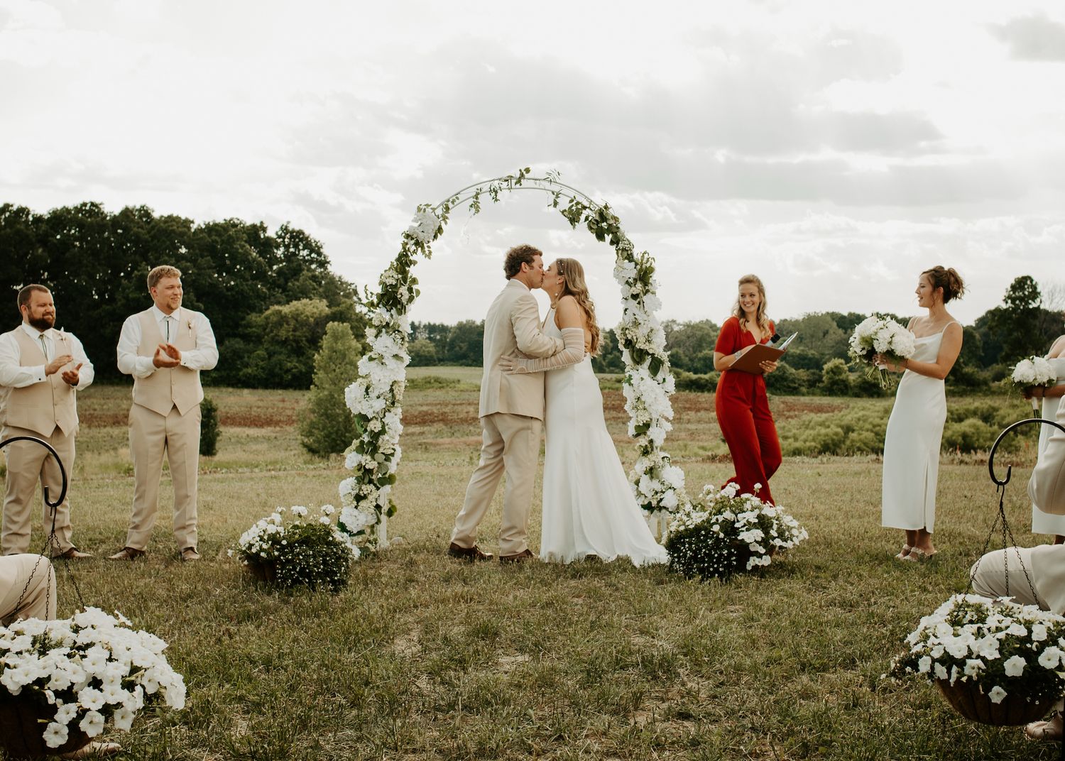 Outdoor wedding ceremony under a floral arch with white flowers and greenery in a scenic field setting.