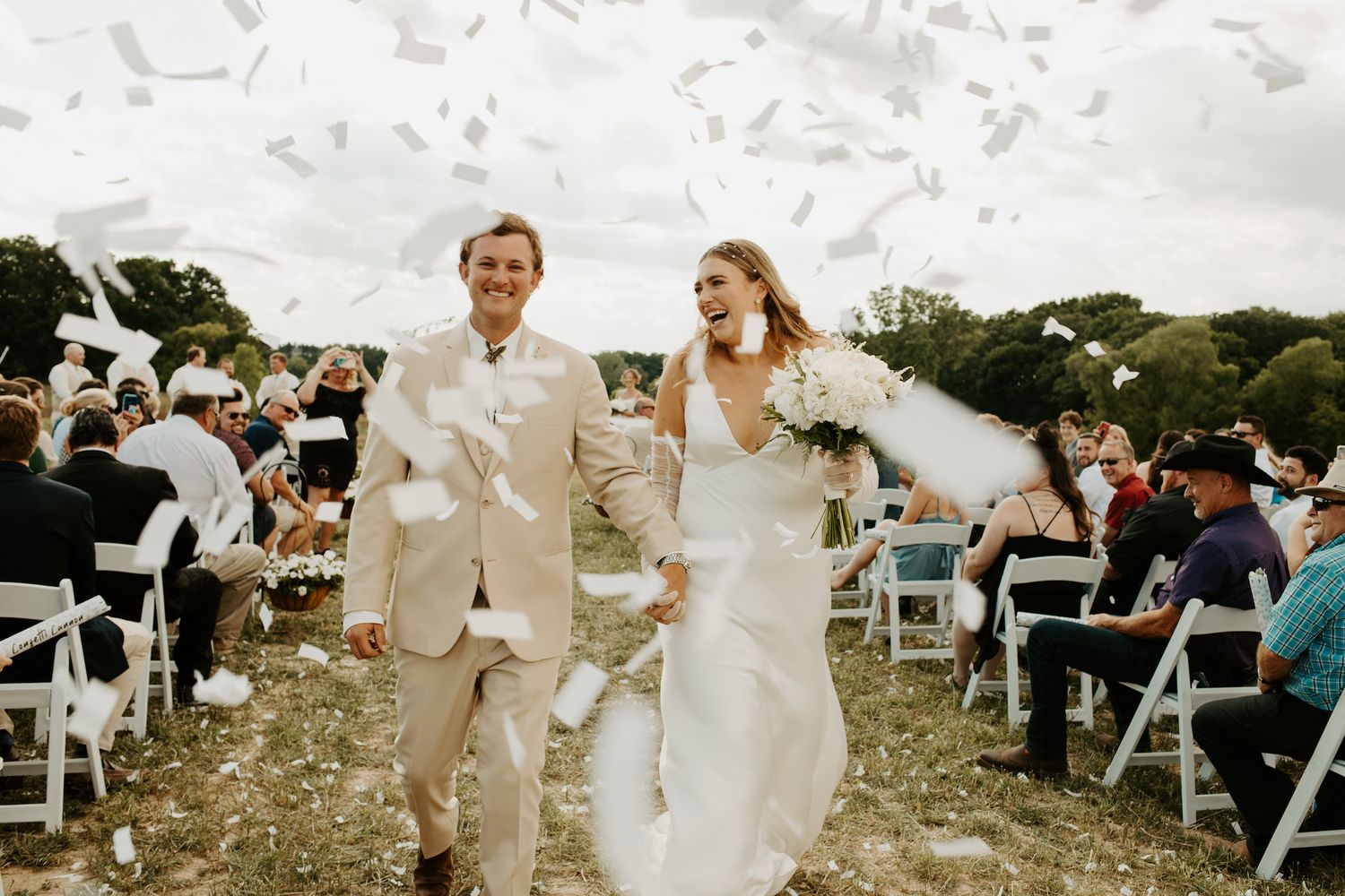 Joyful wedding recessional with confetti being tossed as couple walks down aisle between white chairs.