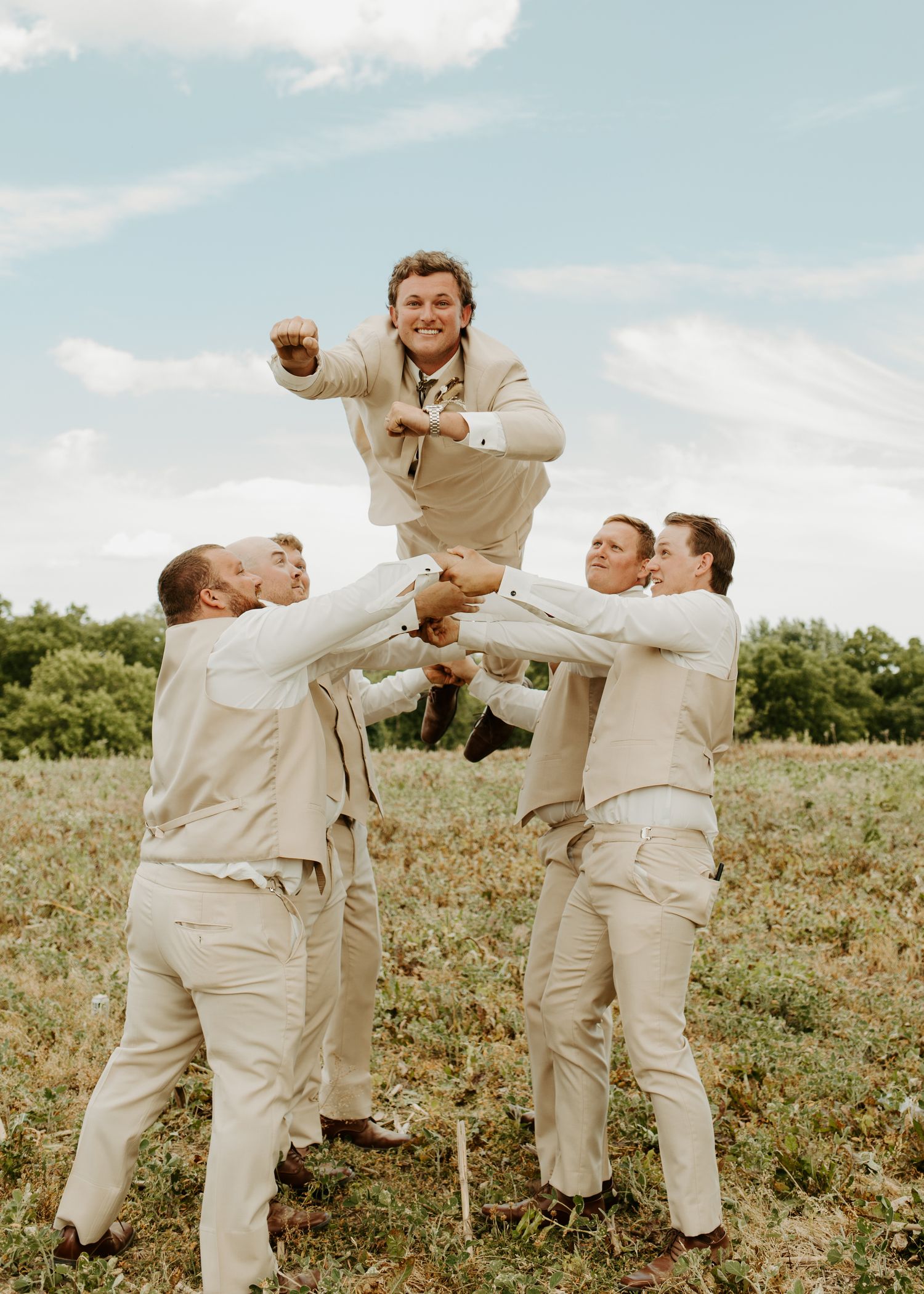 Groomsmen in beige suits playfully lift someone in a coordinated group pose outdoors.