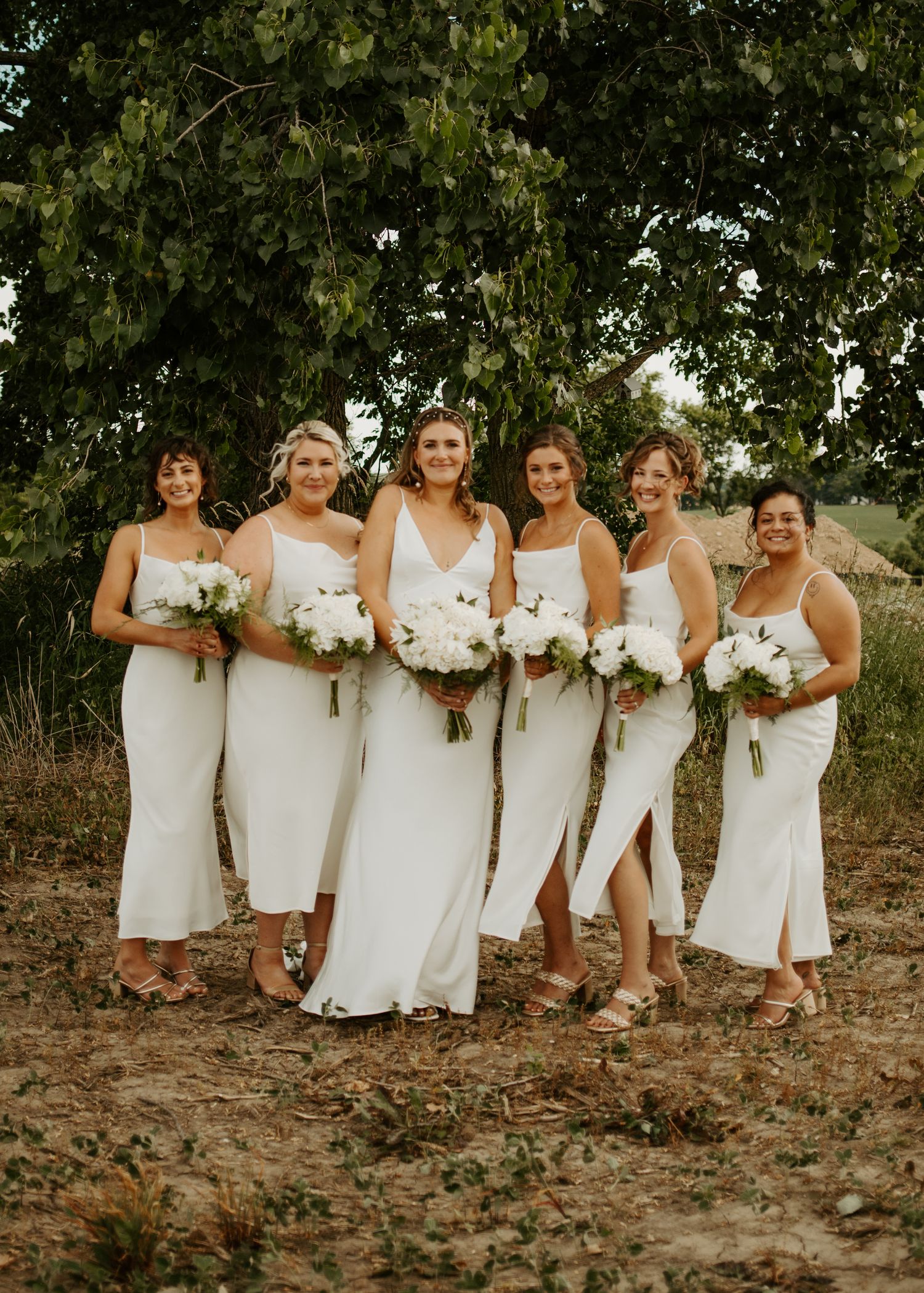 Six attendants in elegant white dresses pose with bouquets against a backdrop of trees.