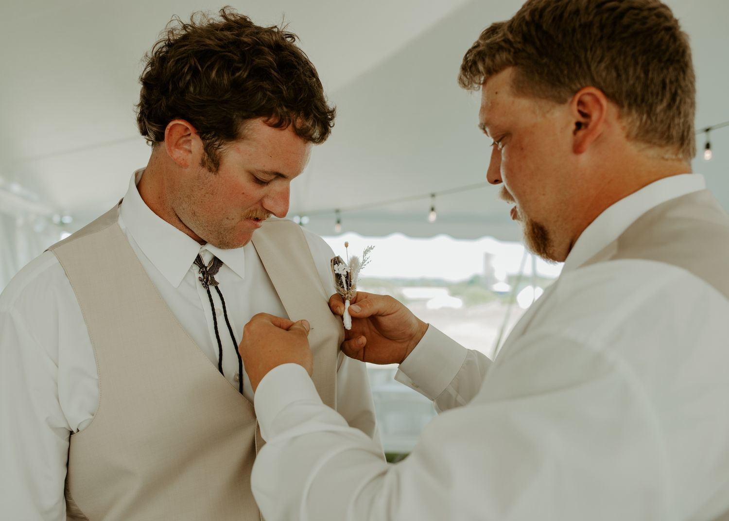 Two people in white formalwear assist with adjusting a tie during wedding preparations.