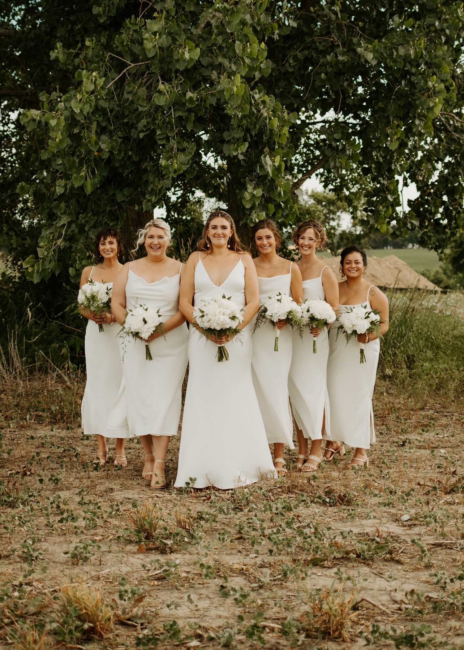 Wedding party members in coordinated white dresses pose with bouquets against natural foliage.
