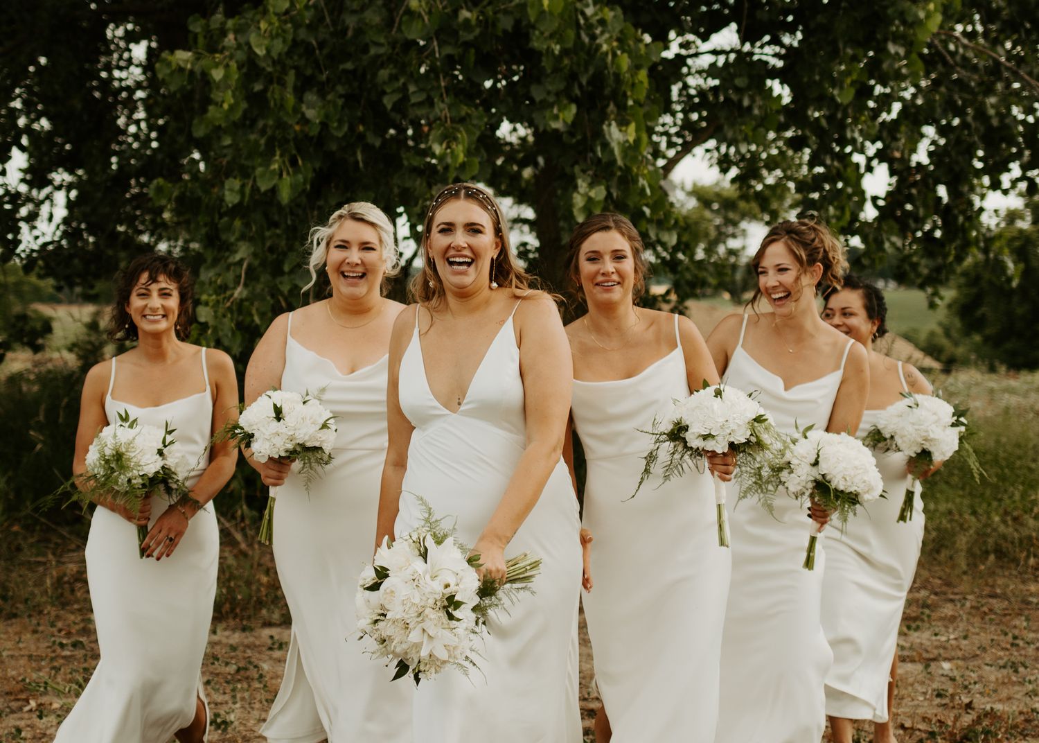 Bridal party in white dresses hold white floral bouquets while posing outdoors.