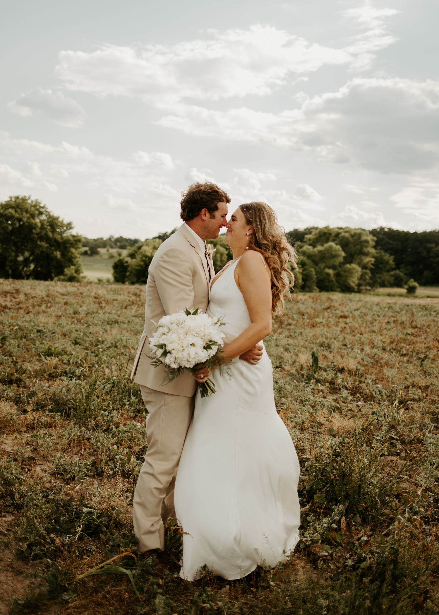 A romantic wedding photo of a couple kissing in a sun-drenched field while holding a white floral bouquet.