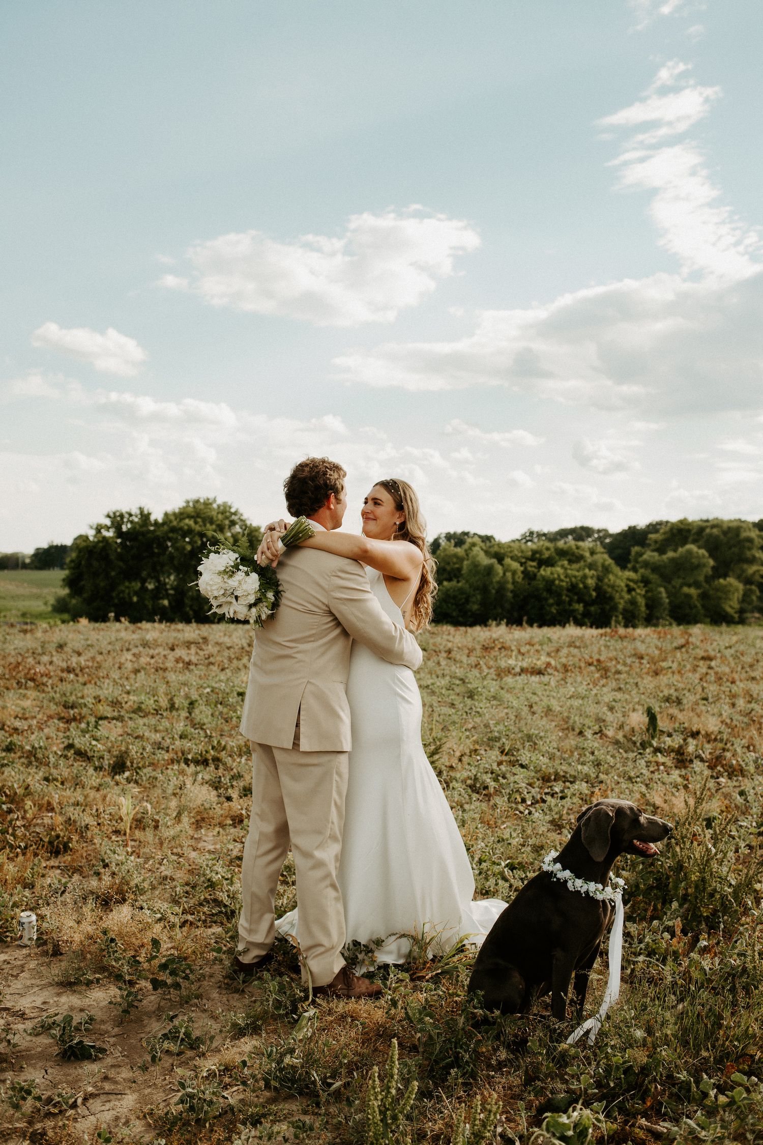 Newlyweds embrace in rustic field setting with their black dog sitting faithfully nearby.