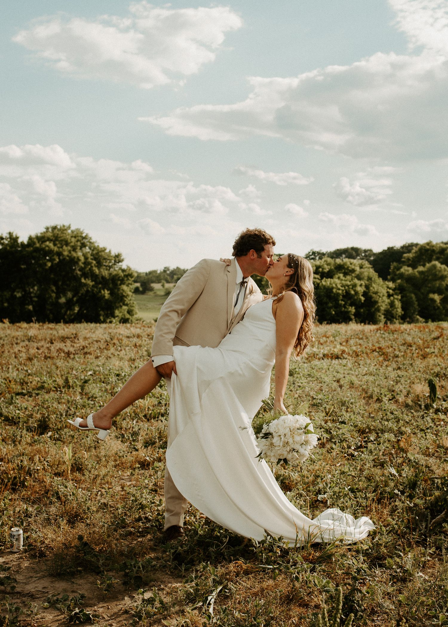 Romantic dip pose between newlyweds in beige and white attire against nature backdrop.