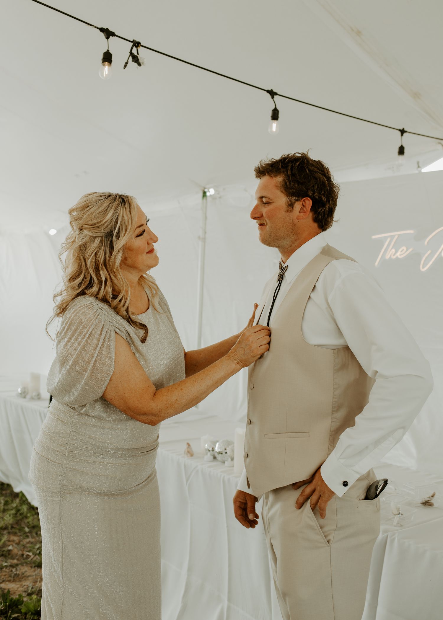Wedding preparation moment in white tent with formal vest and tie adjustments.