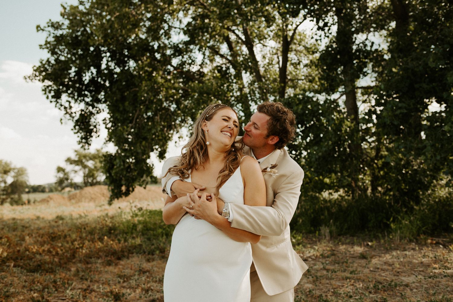 A romantic wedding photo of a couple embracing outdoors under a tree during golden hour.