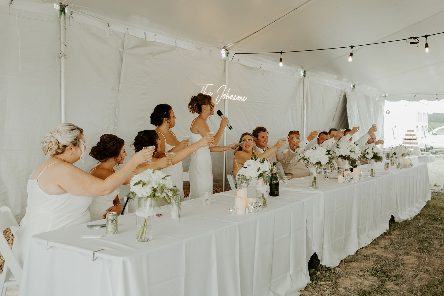 Wedding party seated at an elegant white-draped head table under a tent with floral centerpieces.