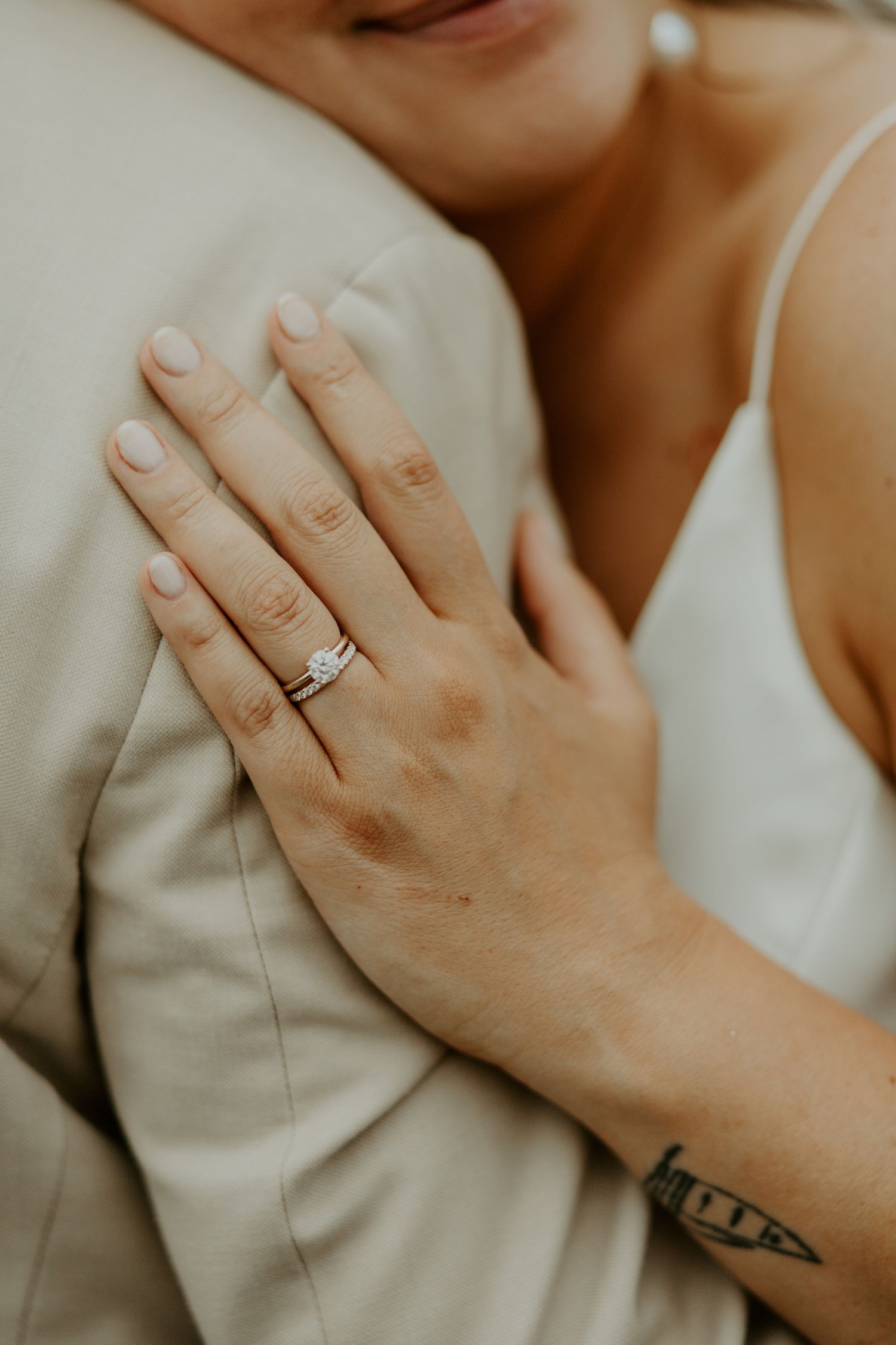 Close-up of hands showing engagement ring and tattoo against beige suit fabric.