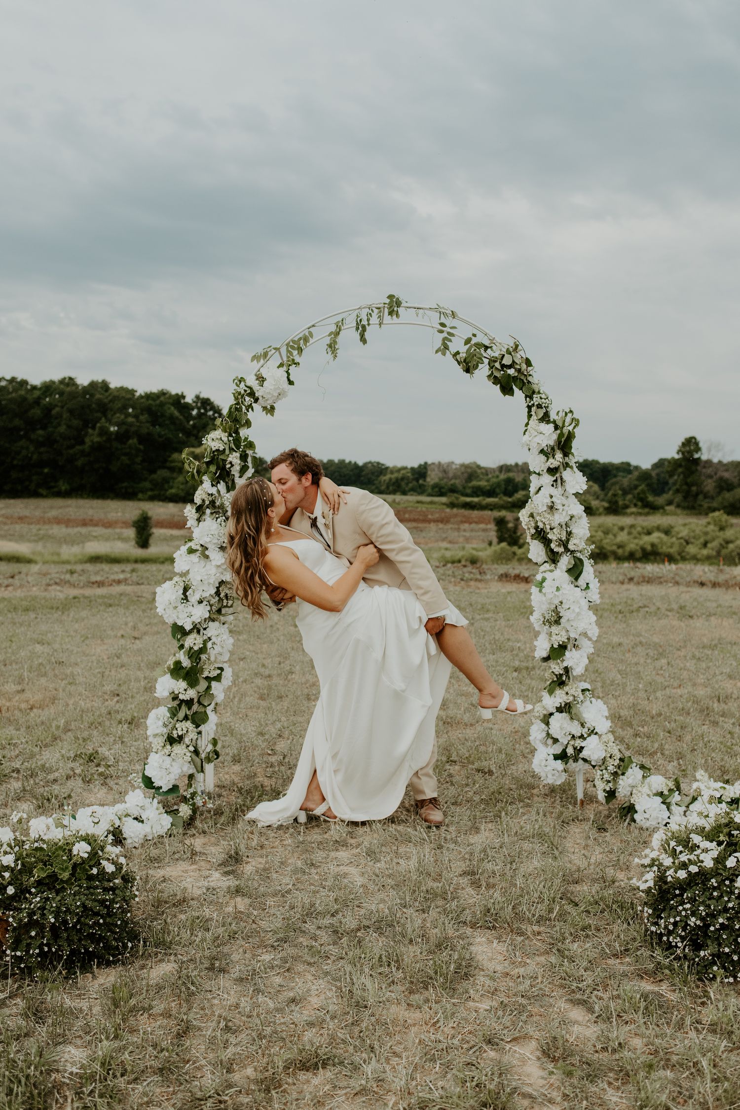 Romantic dip kiss under white floral arch in outdoor wedding setting.